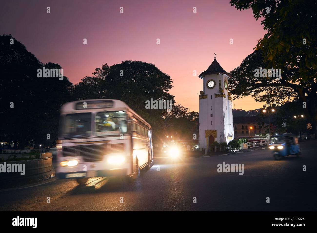 Autobús y tuk tuk en movimiento borroso en el cruce de caminos en la hermosa puesta de sol. Tráfico alrededor de la torre del reloj en la calle en Kandy, Sri Lanka. Foto de stock