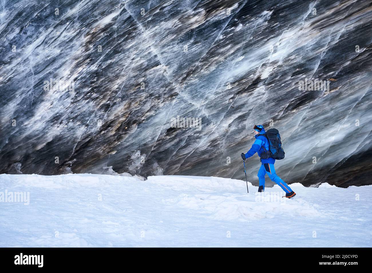 Turista en traje azul con mochila cerca de hielo cueva pared texturizada en glaciar en hermoso paisaje de valle de montaña cubierto de nieve en Almaty, Ka Foto de stock