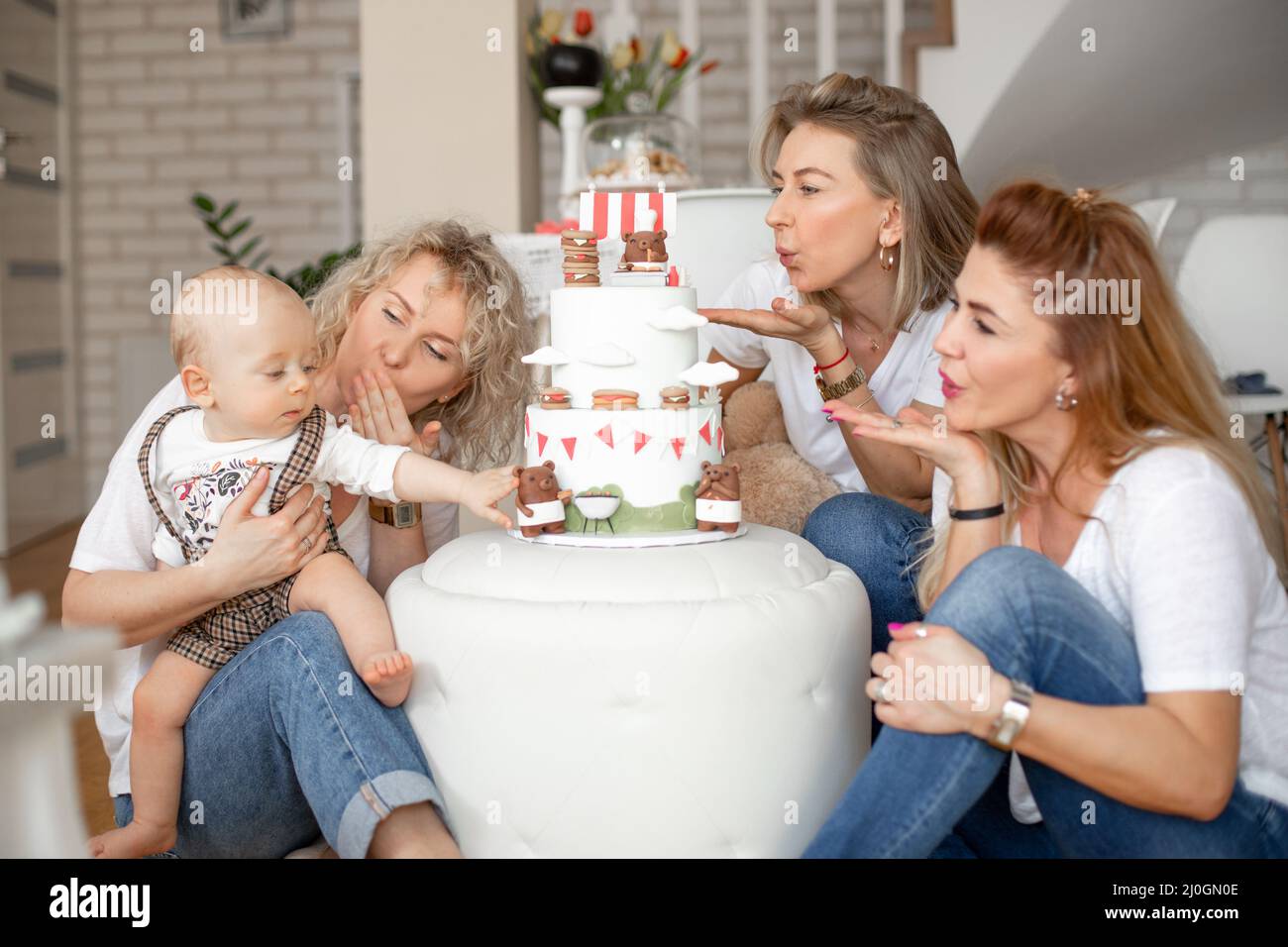 Tres atractivas mujeres de mediana edad y jóvenes que soplan besos al niño de un año tocando el oso en la tarta de cumpleaños. Foto de stock