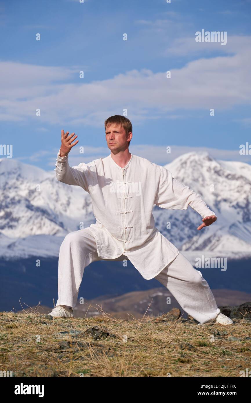 Wushu maestro en un deporte blanco uniforme de entrenamiento kungfu en la naturaleza en el fondo de las montañas nevadas. Foto de stock