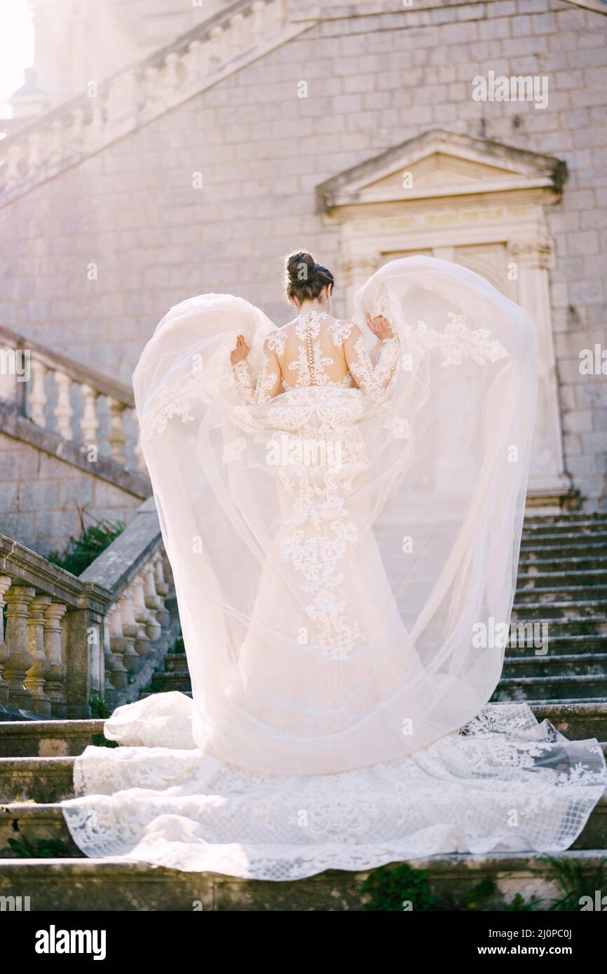 La novia está de pie en los pasos de piedra con sus manos levantando la capa superior de su vestido Foto de stock