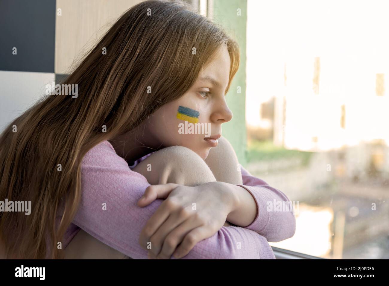 Una triste chica ucraniana con una bandera ucraniana pintada en su mejilla se sienta y mira por la ventana, esperando el final de la guerra. Crisis, guerra, no paz Foto de stock