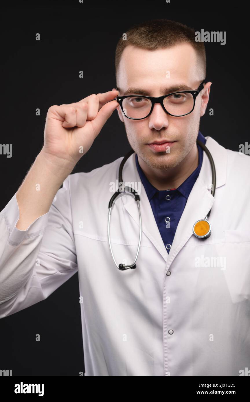 Retrato de un joven médico caucásico en gafas y un abrigo blanco. Sujeta las gafas y mira la cámara de forma tiza Foto de stock