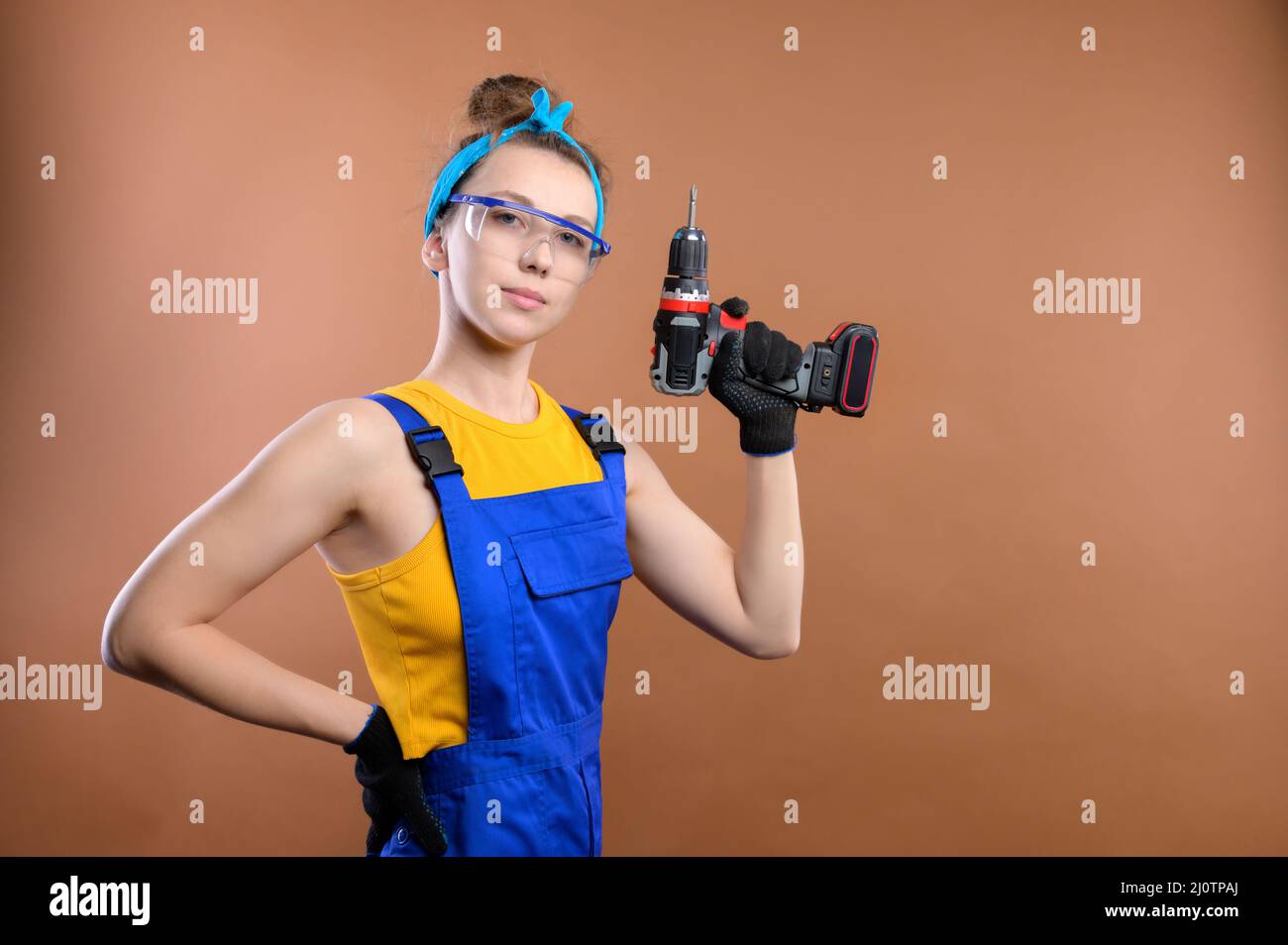 Una joven mujer de handyman muy divertida en una camiseta de tirantes amarilla y monos de trabajo azules con guantes y gafas sobre un fondo marrón claro. Foto de stock