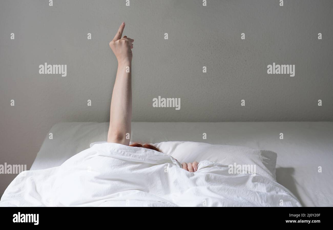 mujer volteando el dedo mientras se esconde debajo de la cubierta de la cama Foto de stock