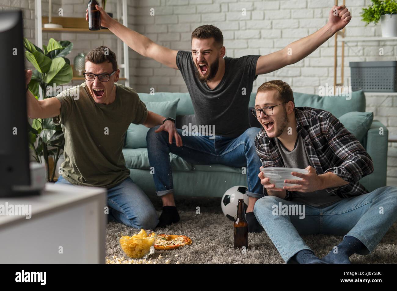 Vista frontal alegre amigos masculinos viendo la televisión deportiva juntos mientras toma aperitivos cerveza. Fotografías de alta calidad Foto de stock
