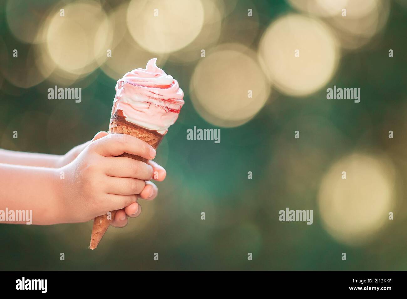 Mano bebé niño sosteniendo un gran helado de fresa en un cono de gofre sobre un fondo bokeh Foto de stock