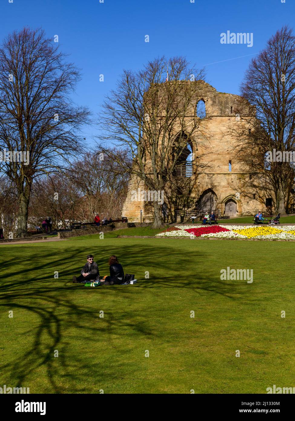 Personas que se sientan relajándose disfrutando del sol (brillantes flores fronterizas, antiguas ruinas de torre, cielo azul) - Castillo de Knaresborough, North Yorkshire, Inglaterra, Reino Unido. Foto de stock