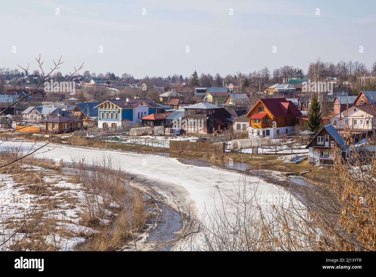 Un río congelado que se extiende en la distancia, un pueblo en la orilla. Primavera, la nieve se derrite, charcos y hierba seca alrededor. Día, tiempo nublado, luz cálida suave. Foto de stock