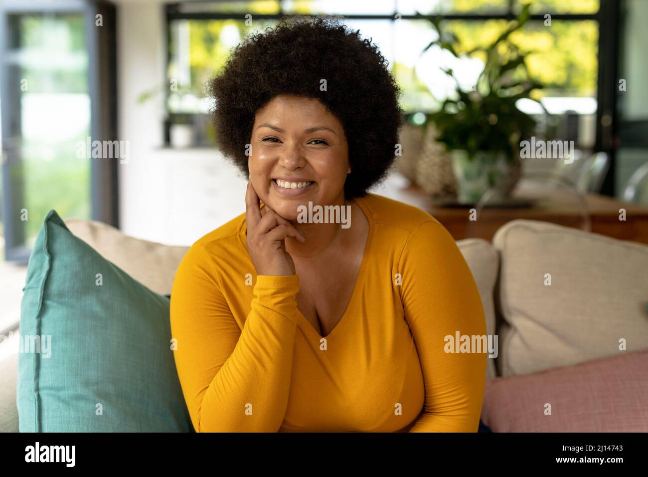 Retrato de una sonriente mujer afroamericana de mediana edad con la mano en la barbilla sentada en el sofá de la casa Foto de stock