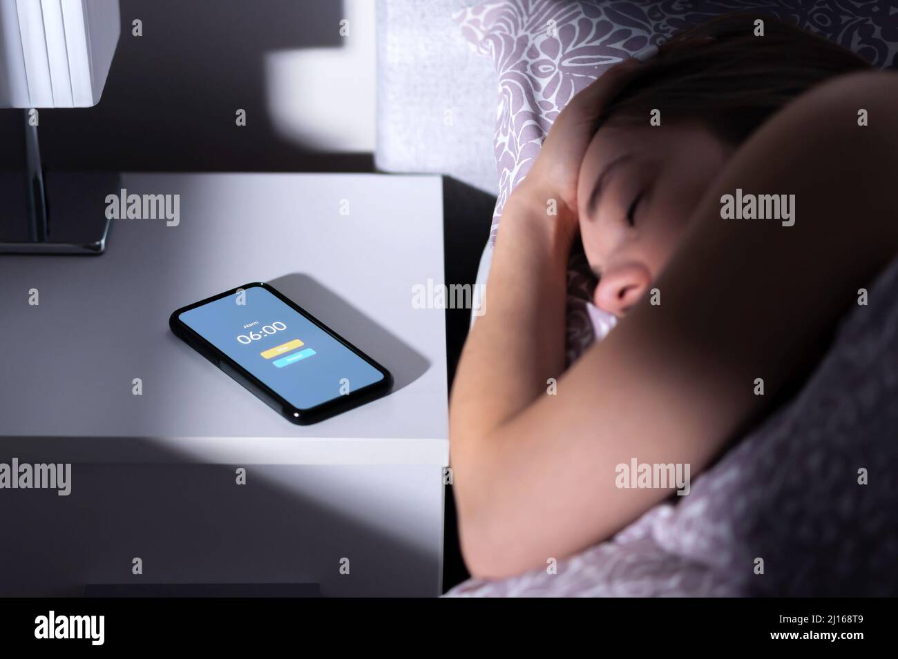 Alarma telefónica despertando a una mujer cansada durmiendo en la cama por la noche o por la mañana. Teléfono móvil en la mesa con temporizador de reloj y botón SNOOZE. Persona que duerme sobre la cama. Foto de stock