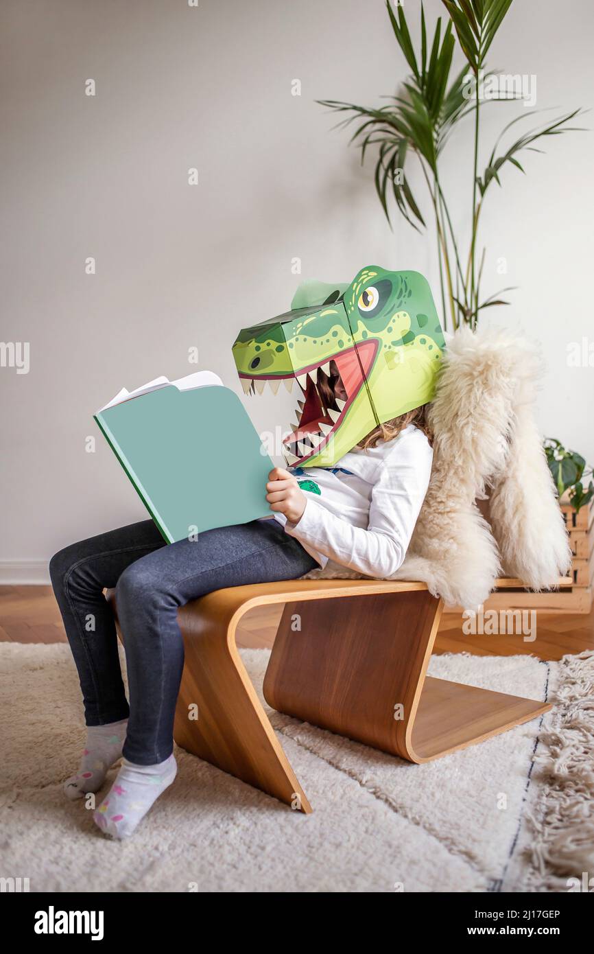 Niña leyendo libro con máscara de disfraces de dinosaurio en casa Foto de stock