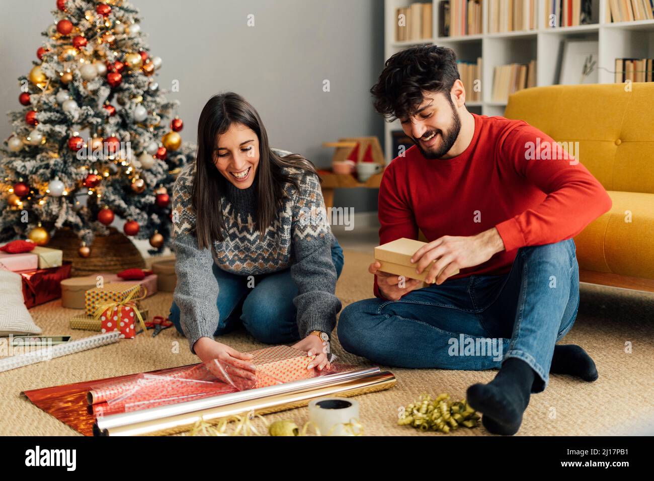 Feliz pareja envolver regalo de Navidad sentado frente al sofá Foto de stock
