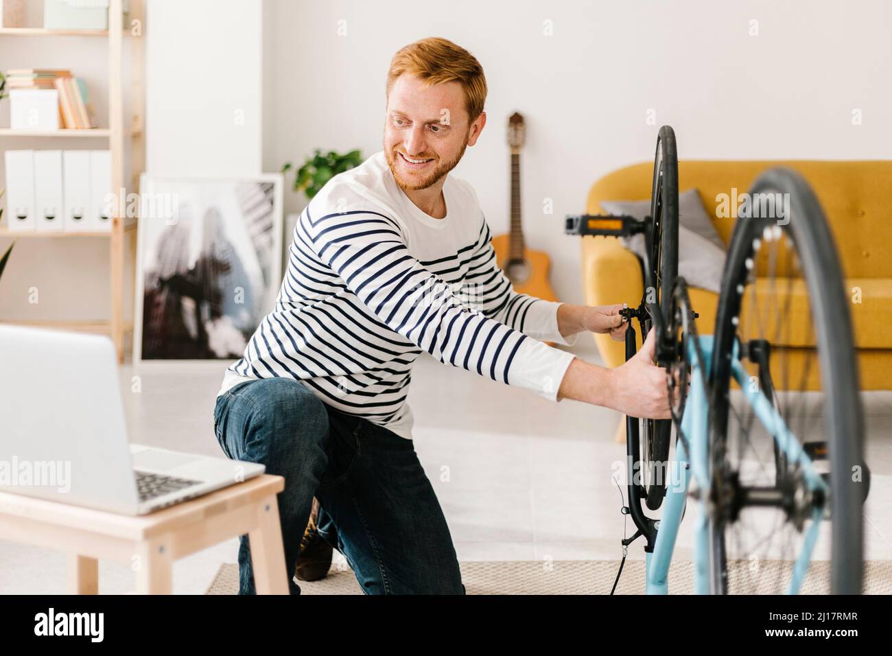 Hombre joven sonriente reparando e-learning en bicicleta a través del portátil en casa Foto de stock