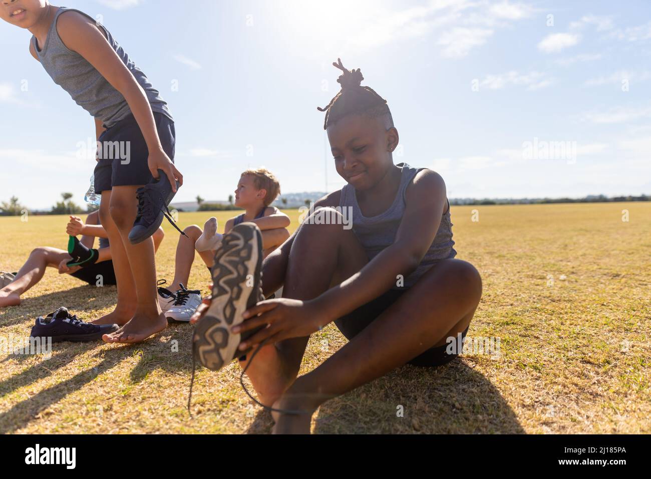 Escolares elementales multirraciales quitando zapatos mientras se sienta en el campo contra el cielo durante el día soleado Foto de stock