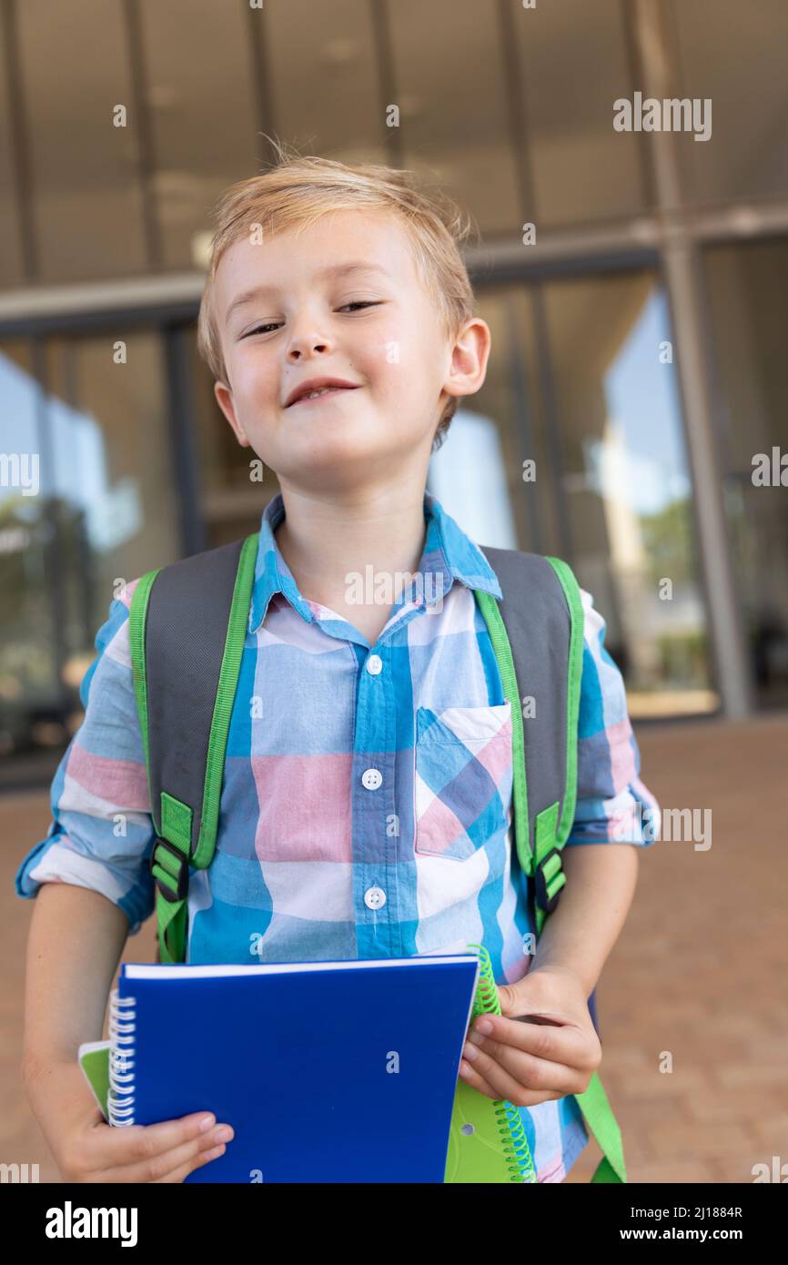 Retrato de un lindo y sonriente niño de escuela primaria caucásico con libros de pie en el campus escolar Foto de stock