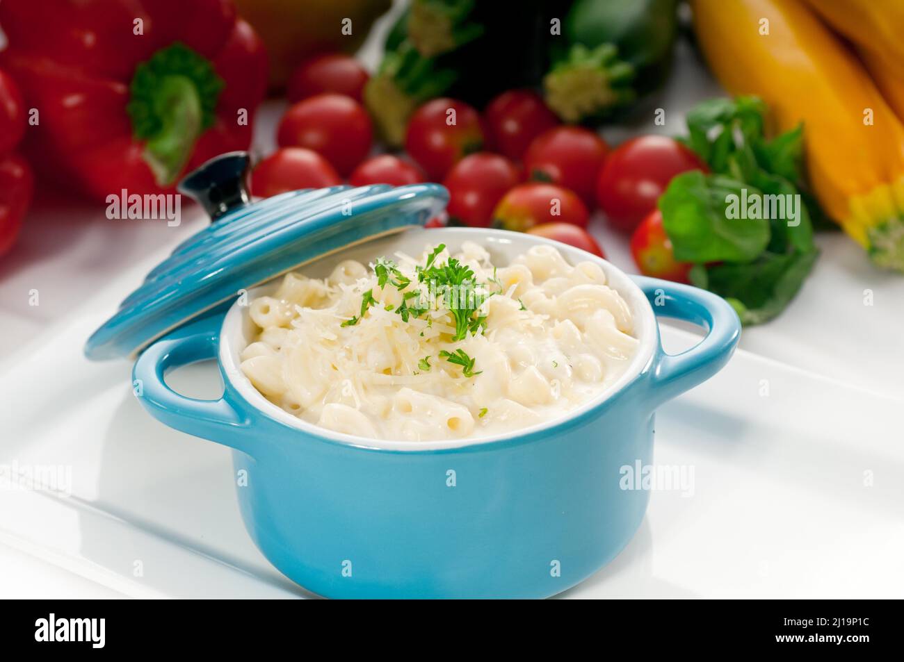 Macarrones y queso, plato favorito de los niños, en una olla de arcilla infantil azul con verduras de fondo Foto de stock