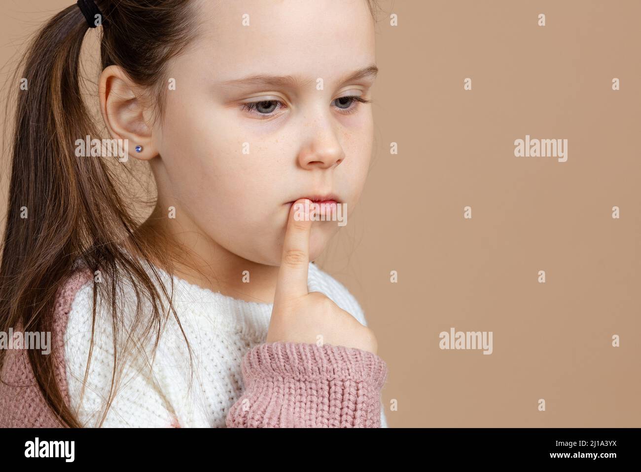 Retrato de joven lindo triste pensivo azul-eyed chica con largo pelo oscuro en blanco y rosa suéter de pie con el dedo índice apuntando a la boca Foto de stock