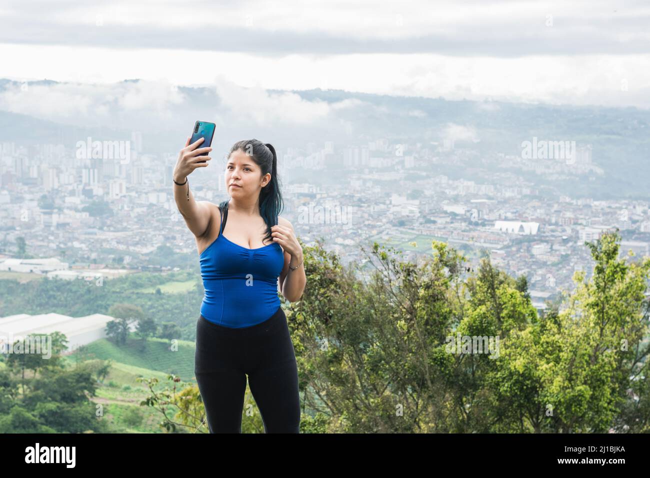 Joven, influencer latino tomando una selfie para sus redes sociales en la cima de una montaña, en el fondo la ciudad de Pereira-Colombia (eje cafete Foto de stock
