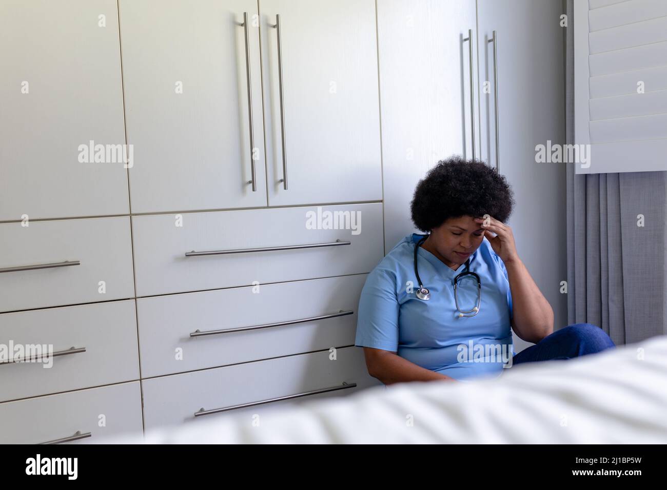 Enfermera de mediana edad afroamericana con la cabeza en la mano sentada en el piso del hospital Foto de stock