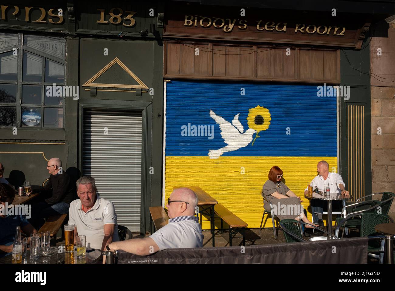 Glasgow, Reino Unido, 25th de marzo de 2022. La gente disfruta de una bebida al sol frente a un mural en la pared de la sala de té de BiddyÕs que representa la bandera ucraniana y una paloma de paz, pintada para mostrar apoyo a Ucrania en su guerra en curso contra la invasión de Rusia, en el extremo este de Glasgow, Escocia, 25 de marzo de 2022. Crédito de la foto: Jeremy Sutton-Hibbert/Alamy Live News. Foto de stock