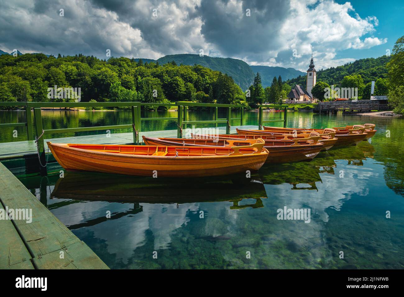 Barcos de madera amarrados en el lago y la iglesia alpina tradicional en el fondo, el lago Bohinj, Ribcev Laz, Eslovenia, Europa Foto de stock