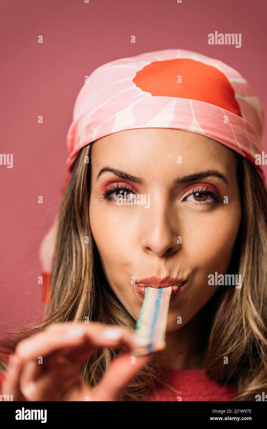 Mujer atractiva con maquillaje comer agrio tira de caramelo colorido y mirar la cámara mientras que de pie sobre fondo rosa en el estudio de luz Foto de stock