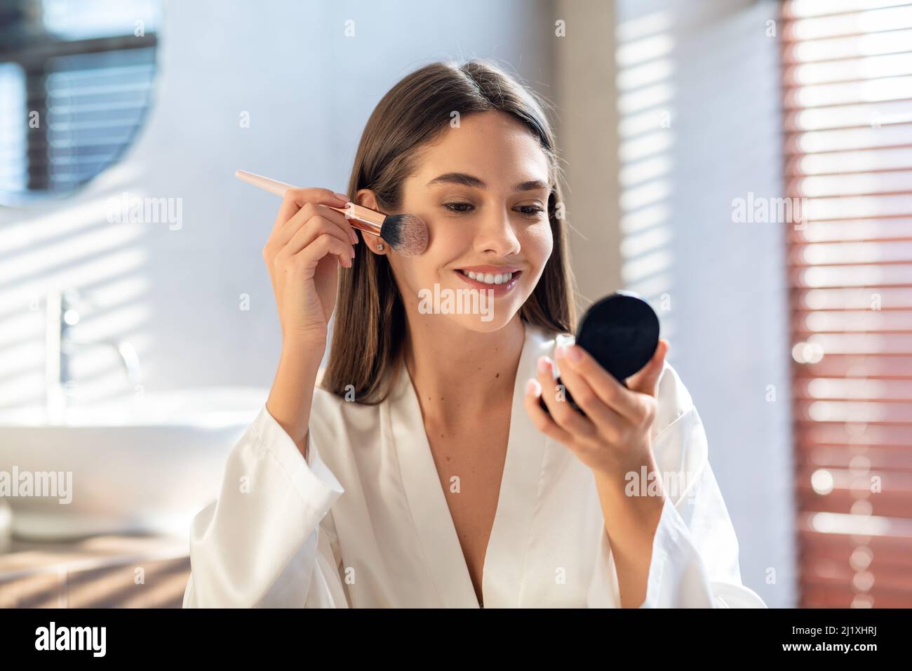 Maquillaje diario. Sonriendo joven mujer aplicando rubor con brocha de maquillaje en el baño Foto de stock