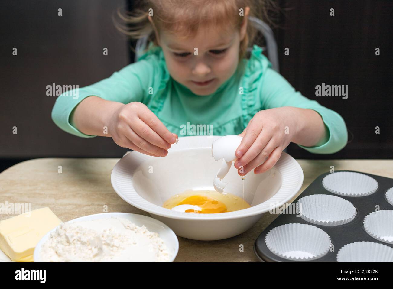 Pequeña niña cocinando en la cocina. Niño sentado en la mesa con comida. Los niños cocinan en casa, aprenden a cocinar muffins, rompiendo el huevo en el tazón. Foto de stock