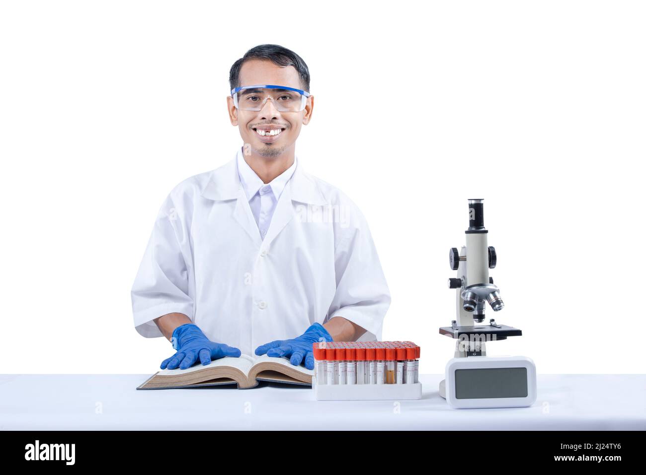 Científico nerd asiático de pie y sosteniendo un libro con un microscopio y una gradilla para tubos médicos en el escritorio aislado sobre fondo blanco Foto de stock