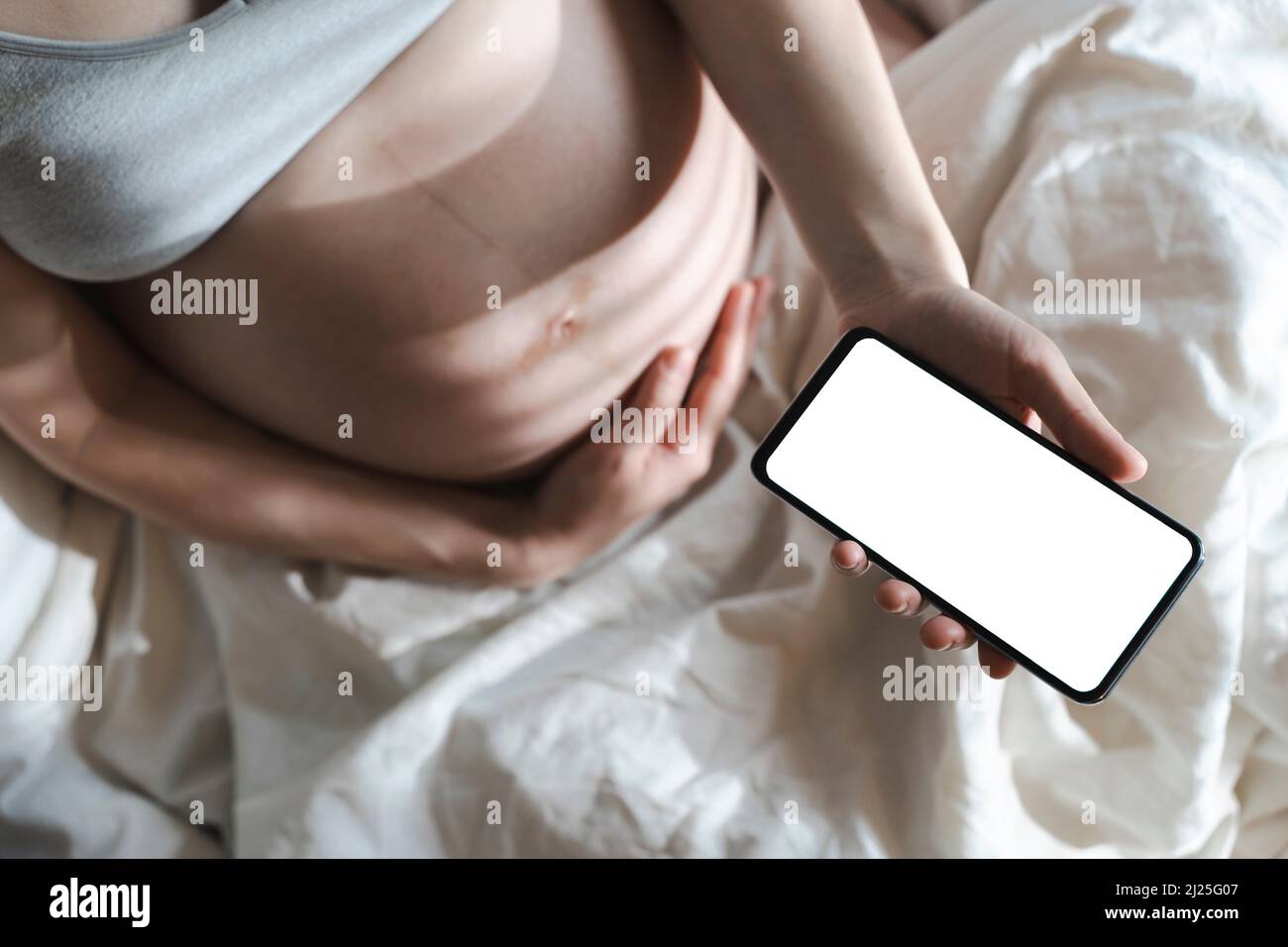 Una mujer embarazada utiliza un smartphone con una pantalla en blanco mientras se relaja en la cama de su casa. Internet, compras, comunicación durante el embarazo y antes del nacimiento de un niño. Salud de las mujeres y los recién nacidos. Fotografías de alta calidad Foto de stock