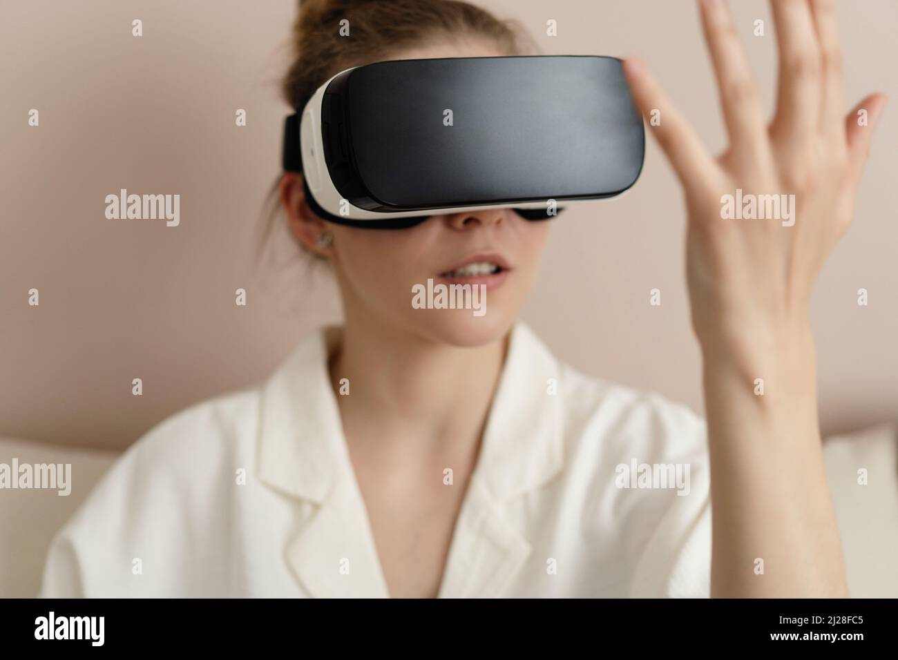 Disfrutar de la nueva realidad. Atractiva mujer joven con auricular VR que gestora y sonríe mientras está sentada en casa Foto de stock