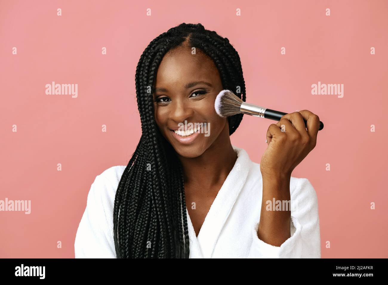 hermosa mujer afroamericana sonriendo aplicando rubor de piel con cepillo de maquillaje sobre fondo rosa, sonriente y feliz Foto de stock