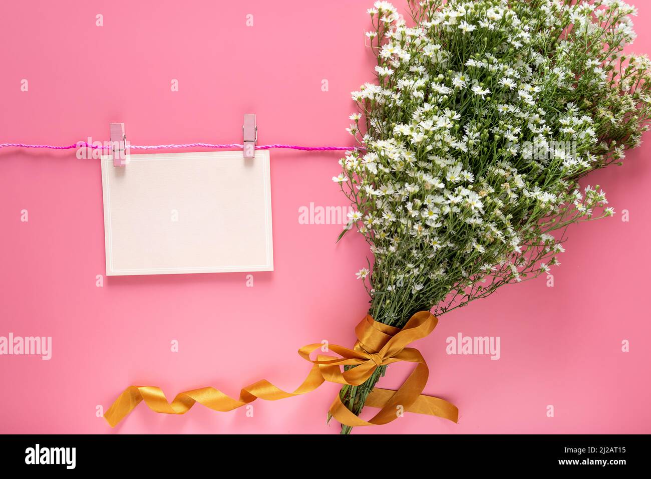 burlete de tarjeta rosa pastel nota hoja de papel en blanco colgar en la cuerda con la clavija de la ropa y primavera fresco blanco ramo de flores aisladas sobre fondo rosa. Foto de stock