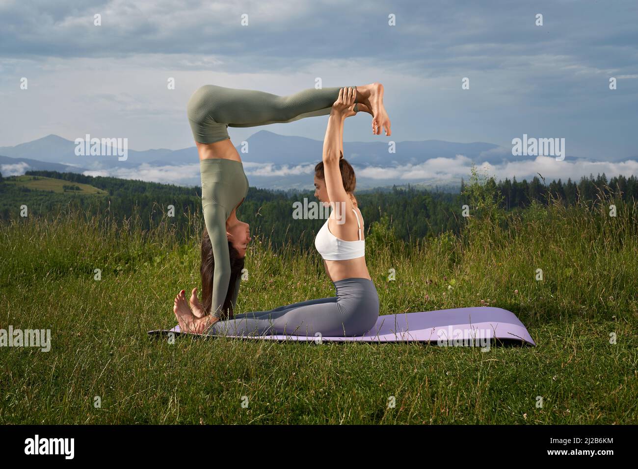 Vista lateral de las mujeres deportivas caucásicas en activo haciendo ejercicio común entre las montañas verdes. Dos mujeres jóvenes haciendo acro yoga durante el día de verano al aire libre. Foto de stock