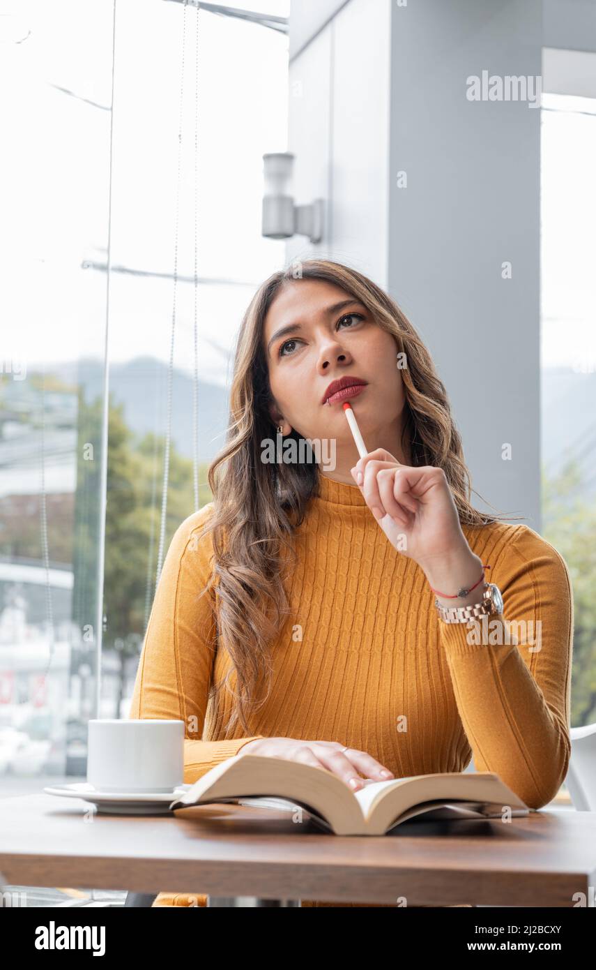 pensive latina joven con maquillaje, sentada con un lápiz, libro abierto y una taza de café en la mesa, joven estudiante estilo de vida y belleza Foto de stock