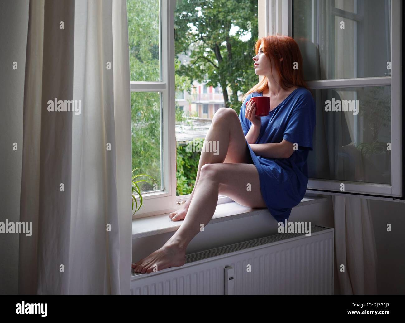 mujer joven con taza de café sentada en el alféizar de la ventana en casa Foto de stock