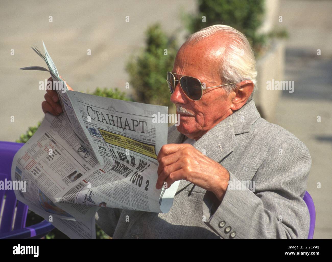 SLAVEIKOV SQUARE, SOFIA, BULGARIA - Un anciano lee el periódico mientras está sentado en la silla en la acera. Foto de stock