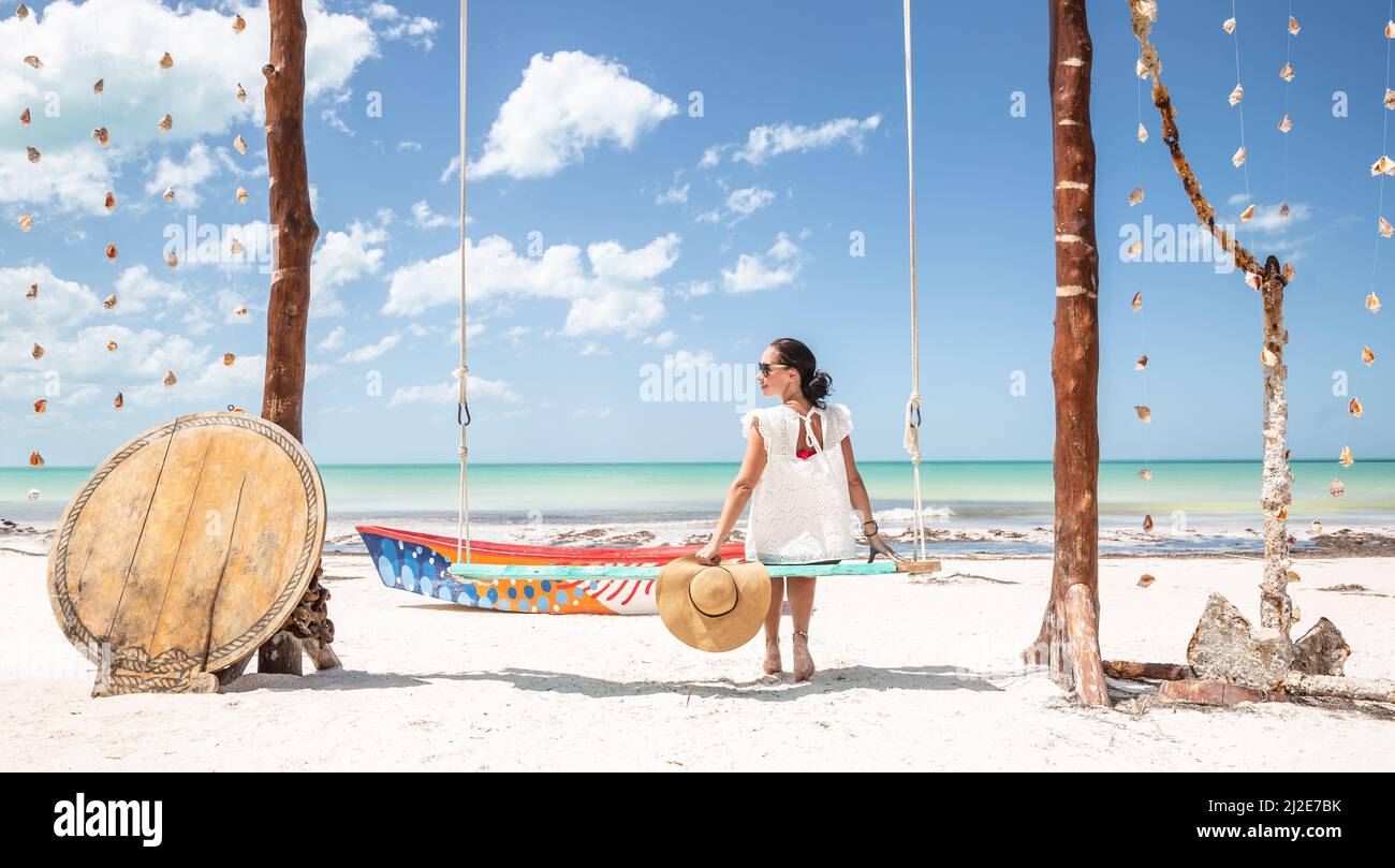 Una mujer joven se sienta en un gran columpio sola en un paraíso tropical durante las vacaciones, sosteniendo sombrero de sol en su mano. Foto de stock