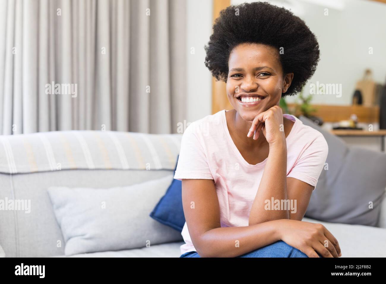 Retrato de una sonriente mujer afroamericana de mediana edad sentada en el sofá con la mano en la barbilla en casa Foto de stock