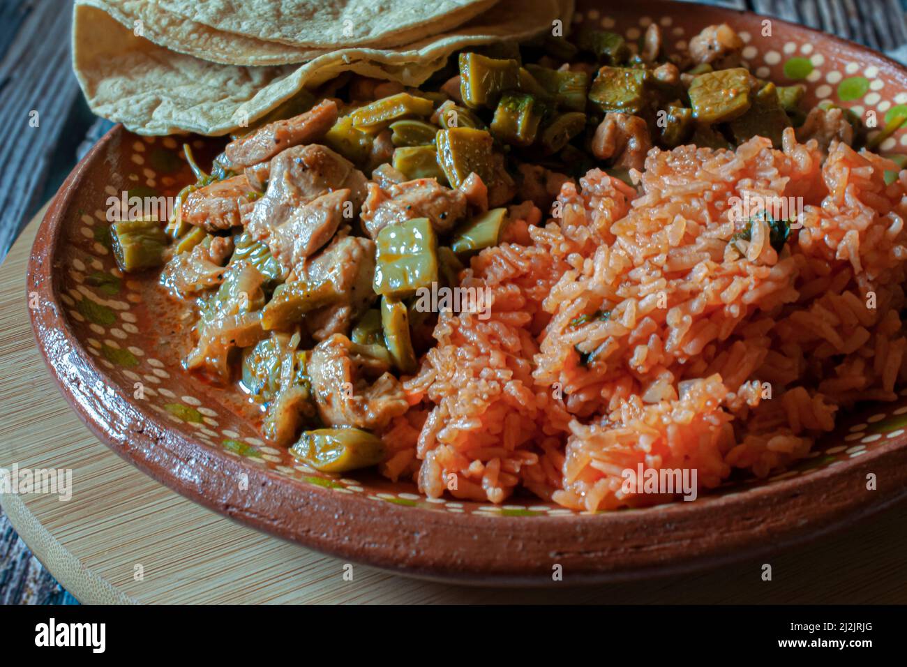 Cerdo. Comida mexicana, pollo con nopales y arroz mexicano con tortillas de maíz hechas a mano en un plato de barro o arcilla, sobre una tabla de corte redonda de madera. Foto de stock