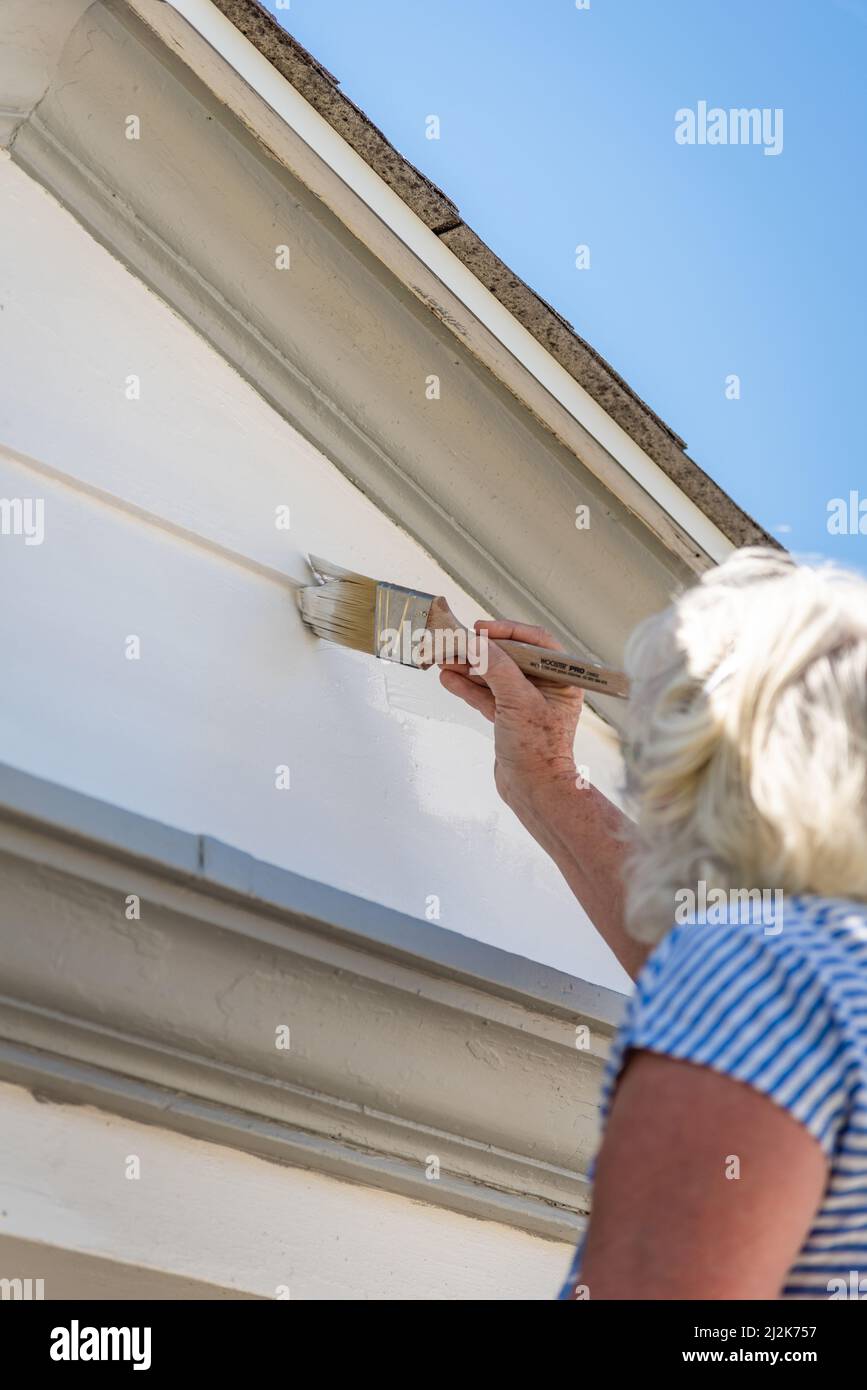 Señora mayor con el pelo blanco pintando un exterior de la casa cerca del techo con pintura y cepillo. Foto de stock