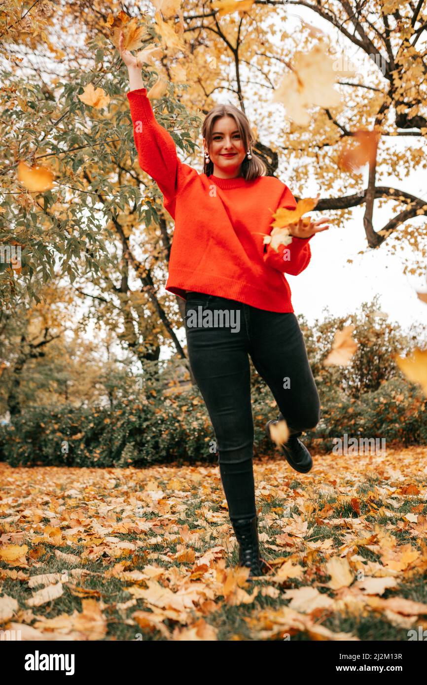 Retrato vertical completo de una joven sonriente saltando una pierna, atrapando hojas de arce del parque naranja de árboles. Foto de stock