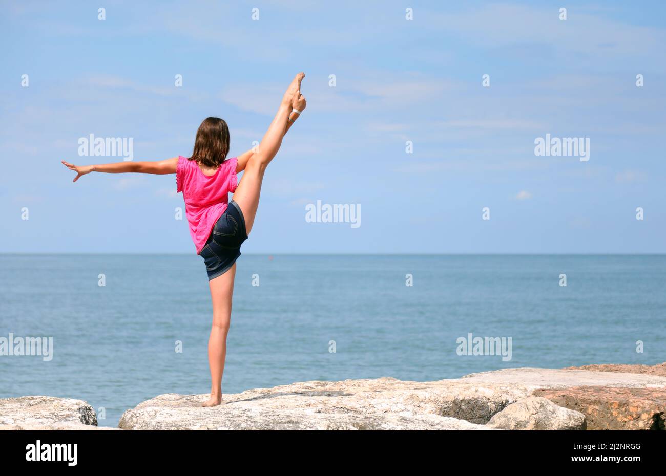 chica joven esbelta con pantalones cortos realiza un ejercicio de gimnasia rítmica su pierna por encima de la cabeza junto al mar el verano de stock - Alamy