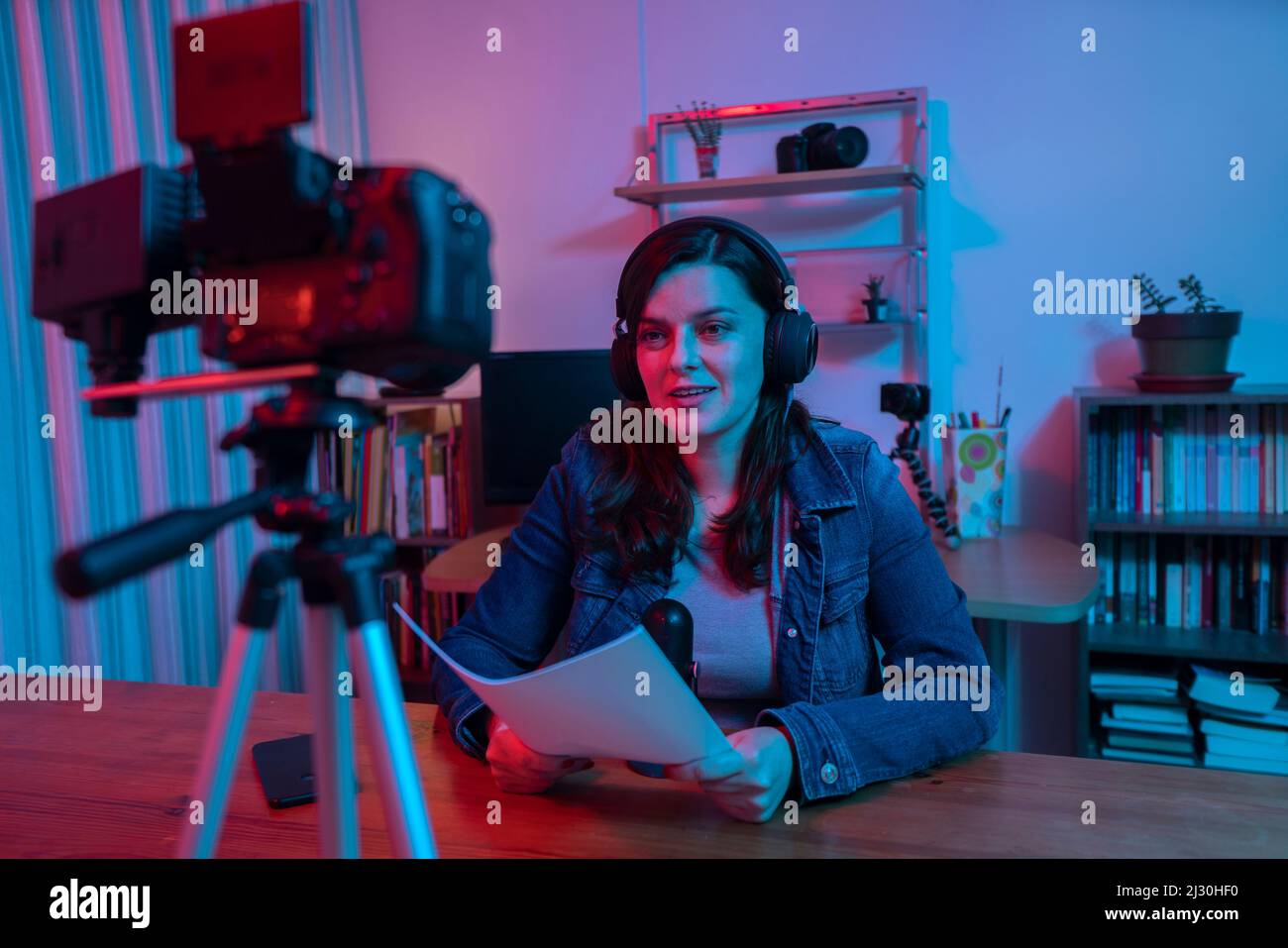 Hermosa mujer hispana frente a una cámara de video grabando un blog en su estudio con luces rojas y azules dentro de su casa. Transmitir una voz en SO Foto de stock
