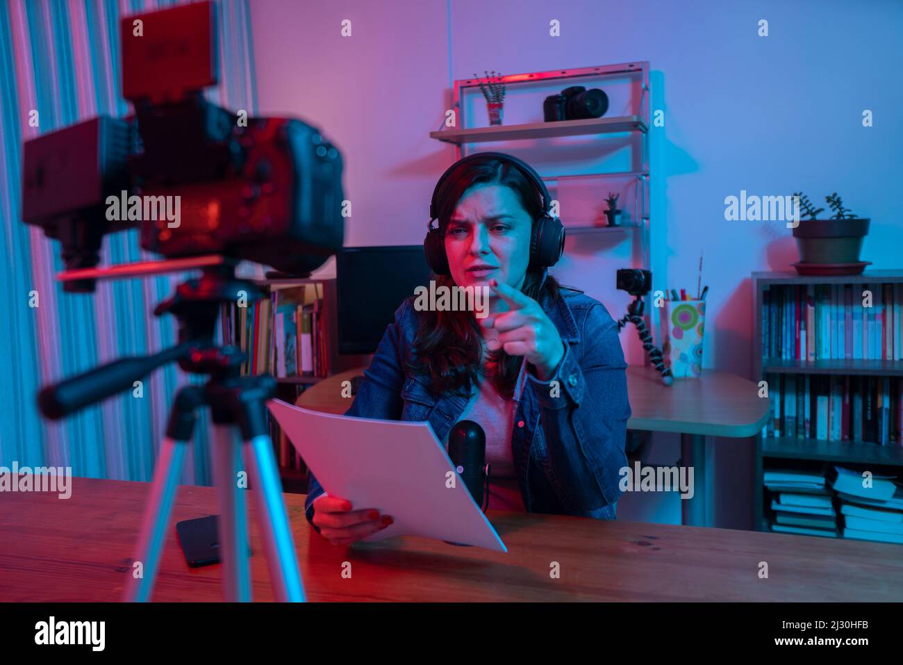 Hermosa mujer hispana frente a una cámara de video grabando un blog en su estudio con luces rojas y azules dentro de su casa. Transmitir una voz en SO Foto de stock