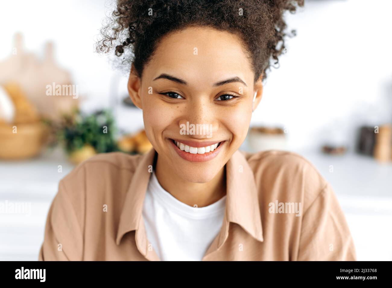 Retrato en primer plano de una encantadora y alegre niña milenaria afroamericana con pelo rizado y pecas, con una sonrisa blanca como la nieve, con una camisa informal, mirando la cámara y sonriendo felizmente Foto de stock