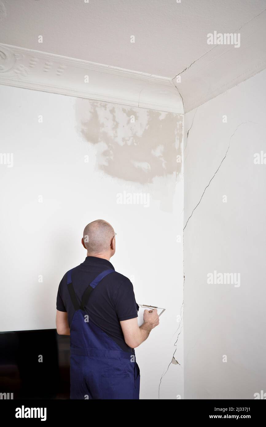 Un portero o portero del edificio inspeccionando una gran mancha de agua y varias grietas en la pared y el techo de la sala de estar de una casa antigua. Foto de stock