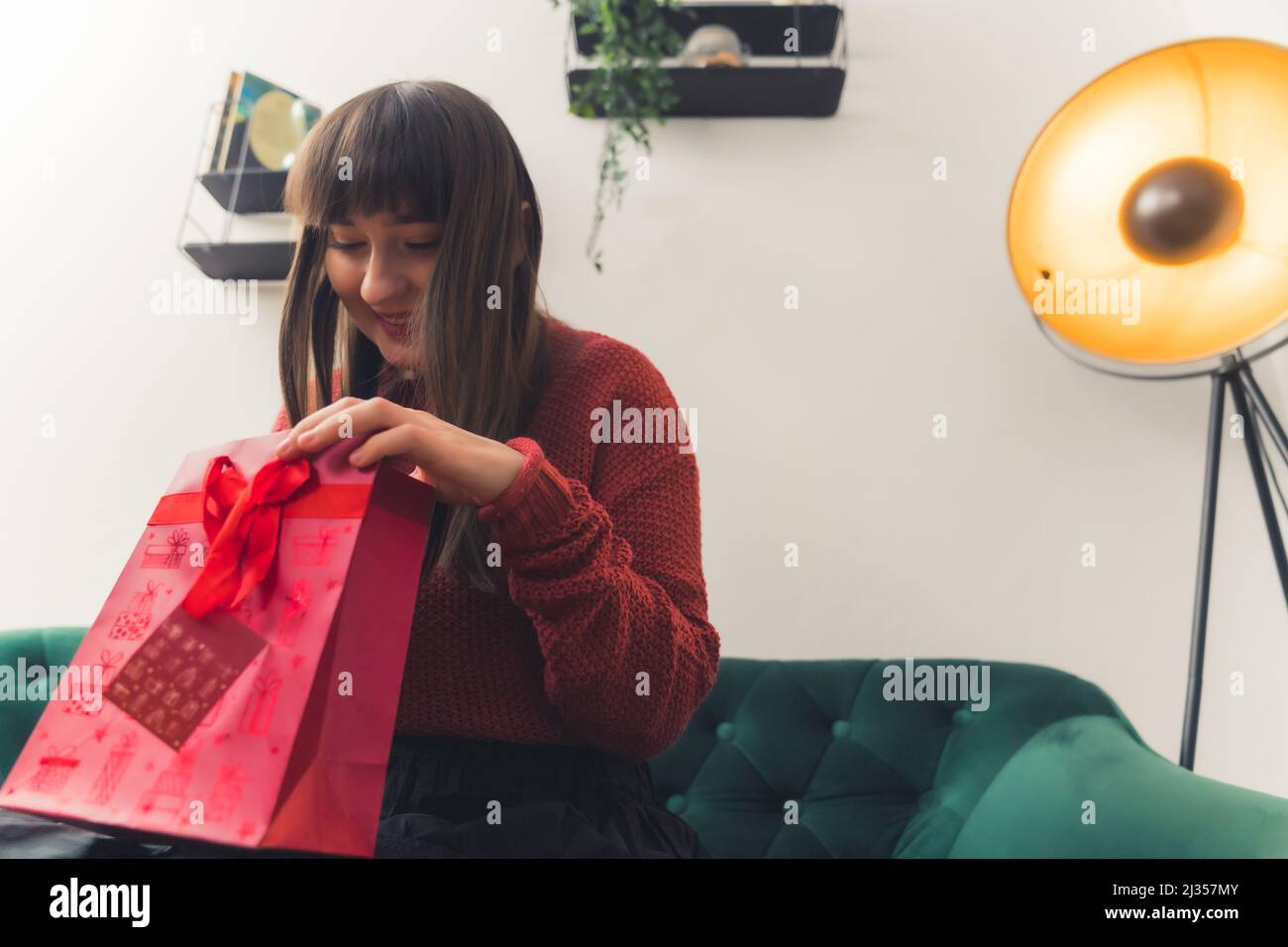Alegre mujer caucásica en sus 20s o 30s abriendo un regalo mientras se sienta en un sofá verde. Disparo en interiores. Fotografías de alta calidad Foto de stock