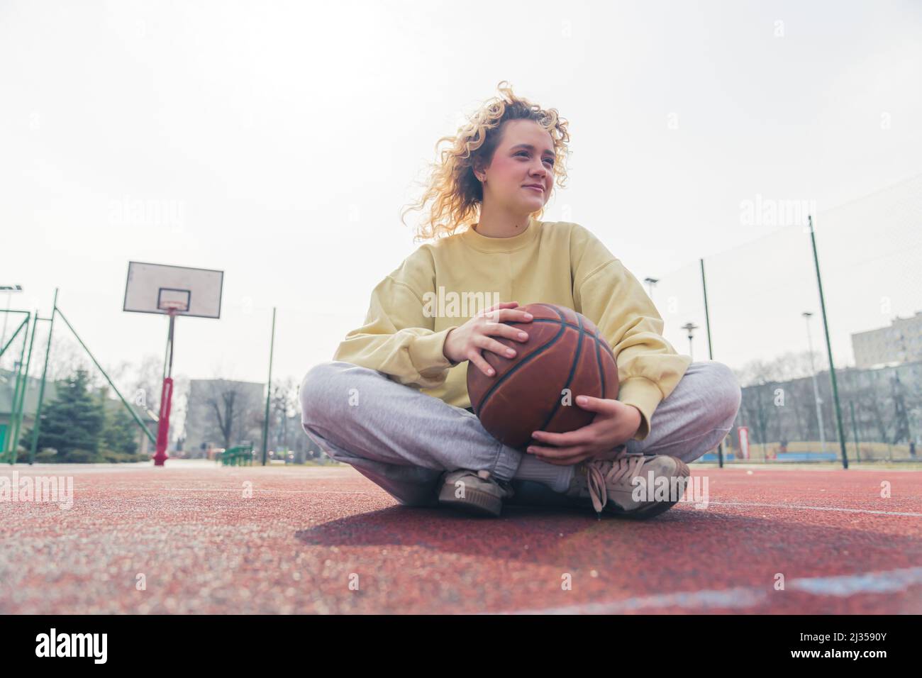 Hermosa joven rubia europea se sienta en la cancha de baloncesto, piernas cruzadas, teniendo pelota de baloncesto en sus manos copia espacio completo tiro . Fotografías de alta calidad Foto de stock